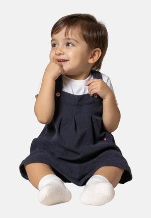Robe salopette en velours côtelé marine avec des accents de boutons, présentant un devant plissé. L'enfant porte un t-shirt blanc et des chaussettes blanches.