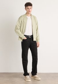 Hellgrüne Bomberjacke mit einem Reißverschluss vorne, getragen über einem weißen T-Shirt. Schwarze Jeans und beige Sneakers runden das Outfit ab. Glatte Stoffstruktur.