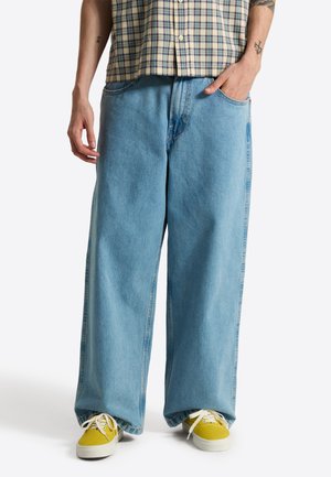 Person trägt hellblaue Jeans mit weitem Bein, gelbe Sneakers mit weißen Schnürsenkeln und ein kariertes Kurzarmhemd, sichtbare Tattoos auf den Armen.