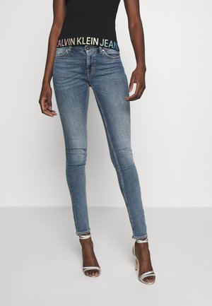 Kobieta ubrana w niebieskie obcisłe dżinsy z wyblakłymi kolanami, czarną bluzkę z wielokolorowym logo Calvin Klein Jeans na wysokości talii oraz białe sandały na wysokim obcasie.