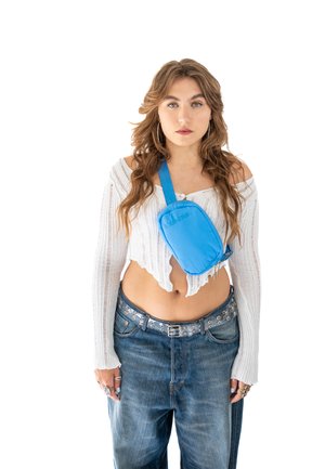 Blue crossbody bag with a smooth, shiny texture and adjustable strap, worn over a white off-the-shoulder top and distressed denim jeans.
