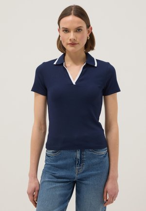 Frau trägt ein marineblaues Polo-Shirt mit kurzen Ärmeln und weißem Besatz, kombiniert mit blauen Jeans, steht vor einem neutralen Hintergrund.