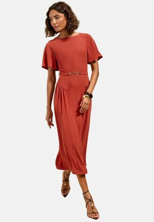 Frau geht, trägt ein rostfarbenes Midi-Kleid mit flatternden Ärmeln, einen Kettengürtel, goldene Armreifen und braune Riemchensandalen.