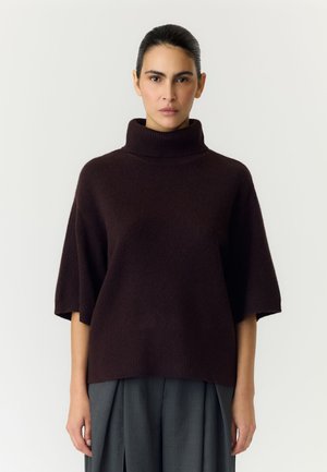 Femme aux cheveux foncés attachés porte un pull à col roulé court marron foncé et un pantalon gris à jambes larges sur un fond uni.