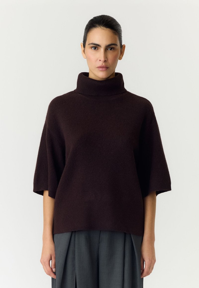 Femme aux cheveux foncés attachés porte un pull à col roulé court marron foncé et un pantalon gris à jambes larges sur un fond uni.