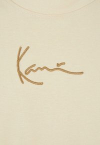 T-shirt crème avec un logo brodé en cursif couleur brun doré à l'avant. Tissu doux et extensible avec un col arrondi.