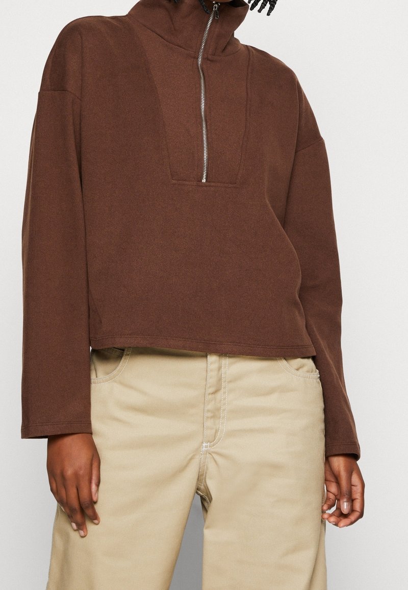 Personne portant un pull-over marron à manches longues avec une fermeture éclair frontale, associé à un pantalon beige, les mains détendues le long du corps.