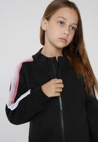 Giacca nera con zip e colletto alto. Presenta strisce multicolori in rosa, blu e bianco sulle maniche. Tessuto dalla superficie liscia.