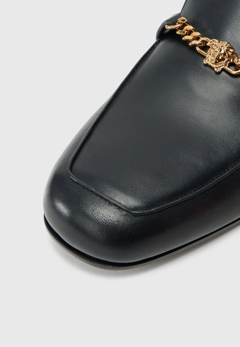 Sort læderloafer med firkantet tå, med en guld kæde-accent og løvehoveddetalje på remmen. Glat tekstur med poleret finish.