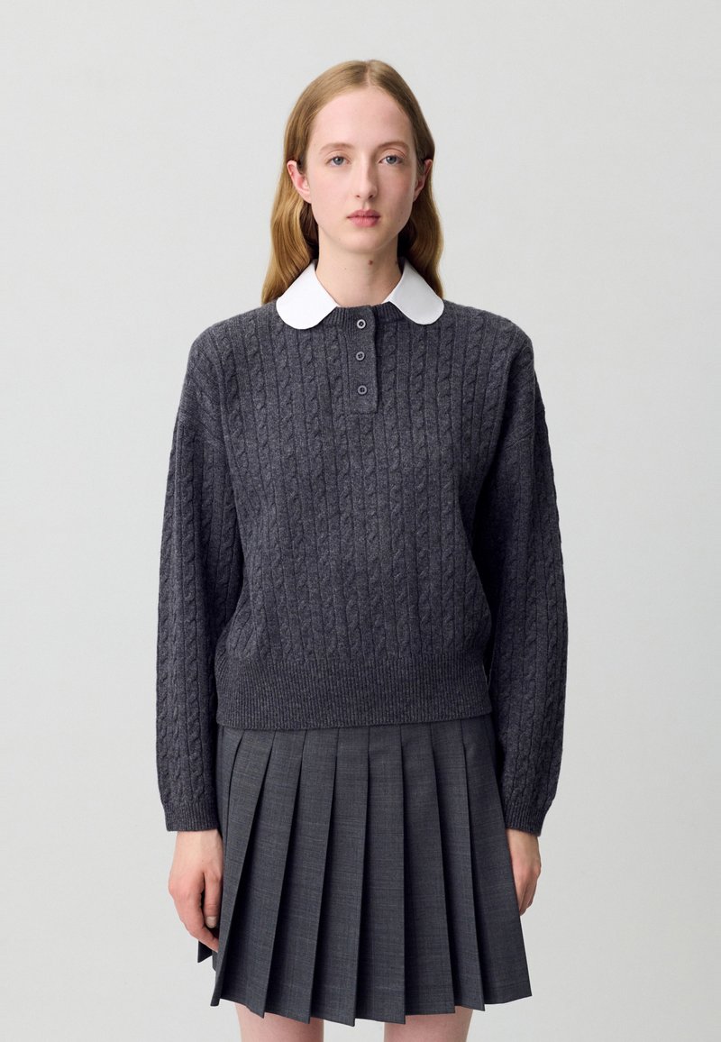 Maglione grigio scuro a maglia rasata con un colletto bianco, con tre bottoni. Abbinato a una gonna grigia plissettata coordinata.