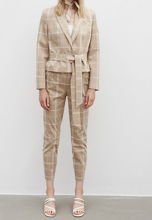 Beige tarkistettu puku, jossa on vyöllinen bleiseri ja kapenevat housut, joissa on valkoiset ruutulinjat. Yhdistetty valkoisiin sandaaleihin.