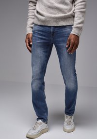 Slim-fit modré džíny s vybledlým vzhledem, spárované s bílými teniskami. Šedý pletený svetr s texturou a vroubkovanými manžetami a lemem.