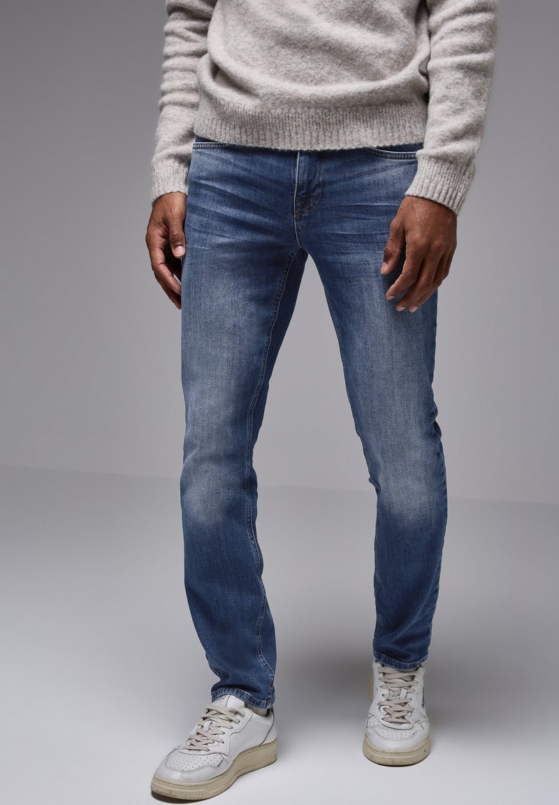 Slim-fit modré džíny s vybledlým vzhledem, spárované s bílými teniskami. Šedý pletený svetr s texturou a vroubkovanými manžetami a lemem.