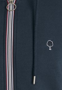 Chaqueta de cremallera navy con cremallera metálica, que presenta un logo de árbol bordado en blanco y un acento a rayas en rojo, blanco y negro.