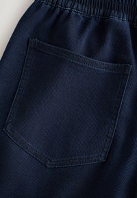 Tissu bleu foncé avec une poche carrée à patche et une taille élastique, montrant des coutures détaillées et une texture.
