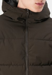 Mørkebrun puffer jakke med lynlås og hætte. Har et tekstureret stof med lodrette striber og justerbare snore.
