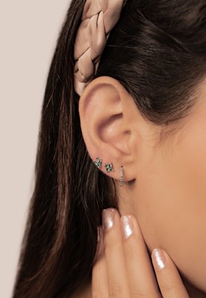 Drei Ohrringe, zwei mit grünen Steinen, einer aus silbernem Metall mit kleinen klaren Steinen, getragen im sichtbaren Ohr neben einer glatten, hellbraunen Haarstruktur.