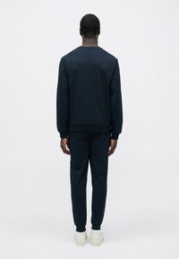 Muž stojící zády, má na sobě tmavě modrou mikinu, odpovídající tepláky, bílé ponožky a bílé tenisky na jednobarevném pozadí.