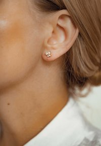 Boucles d'oreilles en ton or, ornées de trois petites perles et d'un accent en cristal, mises en valeur sur une peau lisse, avec des contours d'oreilles visibles.