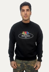 Uomo con capelli scuri che indossa una felpa nera con design floreale colorato e la scritta "PROPAGANDA", e pantaloni con motivo mimetico.