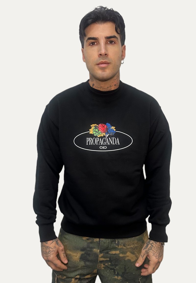 Uomo con capelli scuri che indossa una felpa nera con design floreale colorato e la scritta "PROPAGANDA", e pantaloni con motivo mimetico.