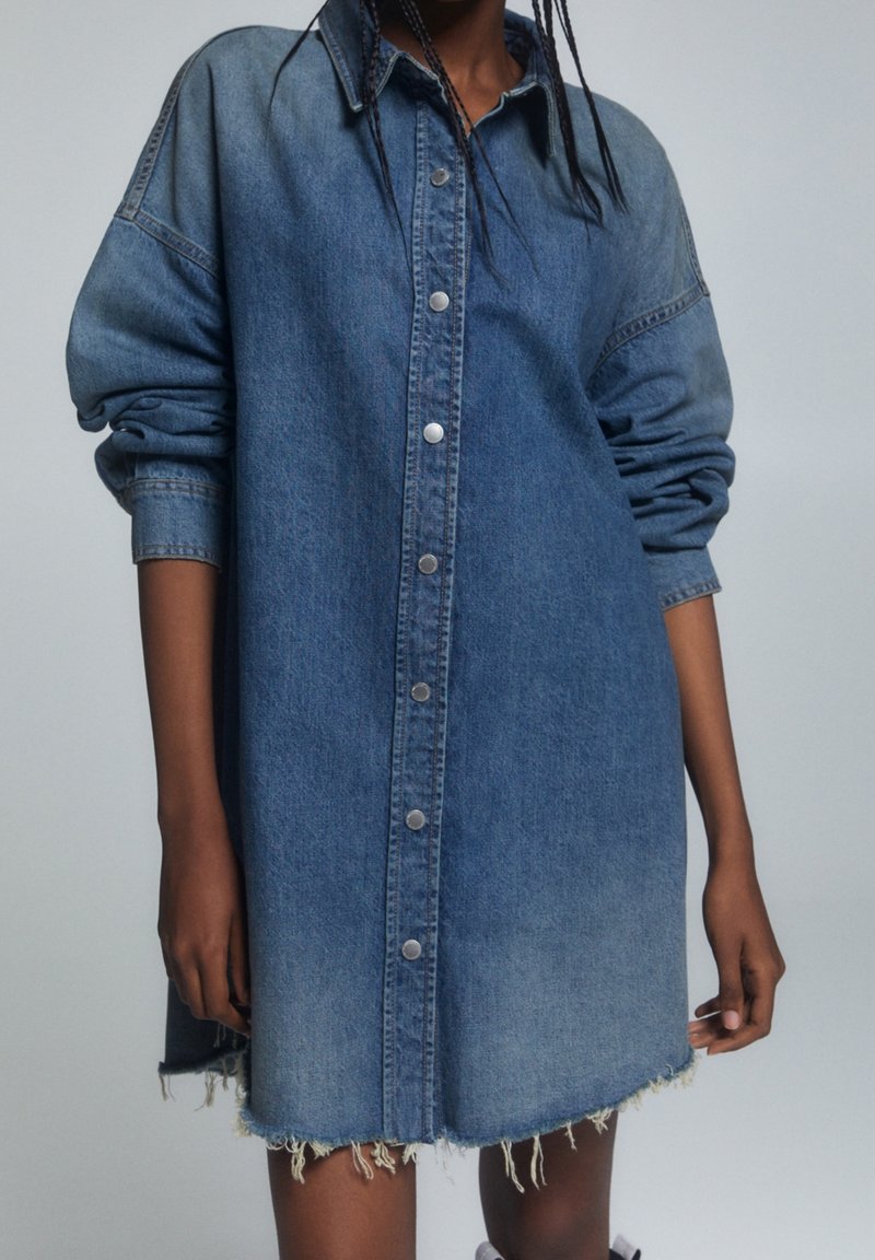 Personne portant une robe en jean bleu à manches longues, boutonnée, avec un ourlet effiloché et des manches retroussées.