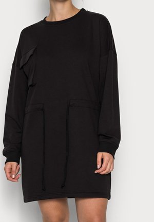 Kvinde iført en sort, langærmet sweatshirt-kjole med snore og lommelapper foran en ensfarvet lys baggrund.