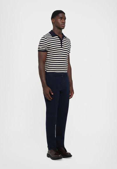 Marineblau-weiß gestreiftes Polohemd mit Kragen, kombiniert mit dunkelblauen Hosen und braunen Schnürschuhen. Glatte Textur, taillierte Passform.