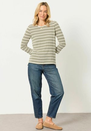 Lyshåret kvinde iført en langærmet, stribet skjorte, blå jeans og beige slip-on sko, stående med hænderne bag ryggen.