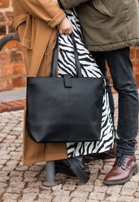 Zwarte leren draagtas met twee lange handvaten, een gladde textuur, minimaal ontwerp, vastgehouden naast een zwart-wit tas met zebra-patroon.