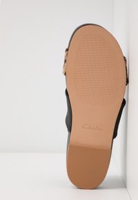 Semelle de sandale avec grip texturé marron clair, bords noirs et brides à motif léopard, positionnée verticalement contre un coin blanc.