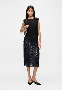 Femme portant un haut noir transparent sans manches, une jupe midi noire à sequins, des talons pointus noirs, tenant un petit sac à main imprimé léopard sur l'épaule.