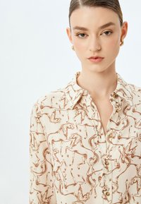 Kraagblouse van crèmekleurige stof met een bruin paardenpatroon, voorzien van knopen aan de voorkant en een gladde, lichte textuur.