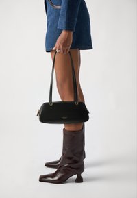 Black leather handbag with a curved shape and gold hardware, held by a hand. Model wears brown, glossy knee-high boots with a chunky heel.