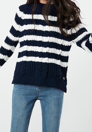 Persona vistiendo un suéter de punto a rayas azul marino y blanco con patrones texturizados, combinado con jeans de mezclilla azul claro.