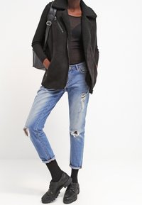 Donna che indossa un gilet in shearling nero, una maglietta nera a maniche lunghe, jeans blu strappati, calze nere, scarpe nere e che porta uno zaino nero.