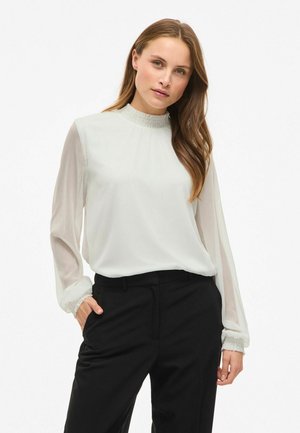 Blouse légère et fluide blanche avec un col haut et des détails froncés. Dotée de longues manches transparentes avec des poignets élastiques. Associée à un pantalon noir.