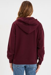 Felpa con cappuccio bordeaux dal texture morbido, con spalle scese e polsini a coste, abbinata a jeans di denim azzurro chiaro.