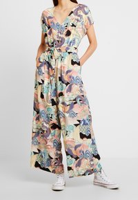 Woman wearing a colorful wide-leg jumpsuit with belt, featuring abstract bird and wave patterns, paired with white high-top sneakers.