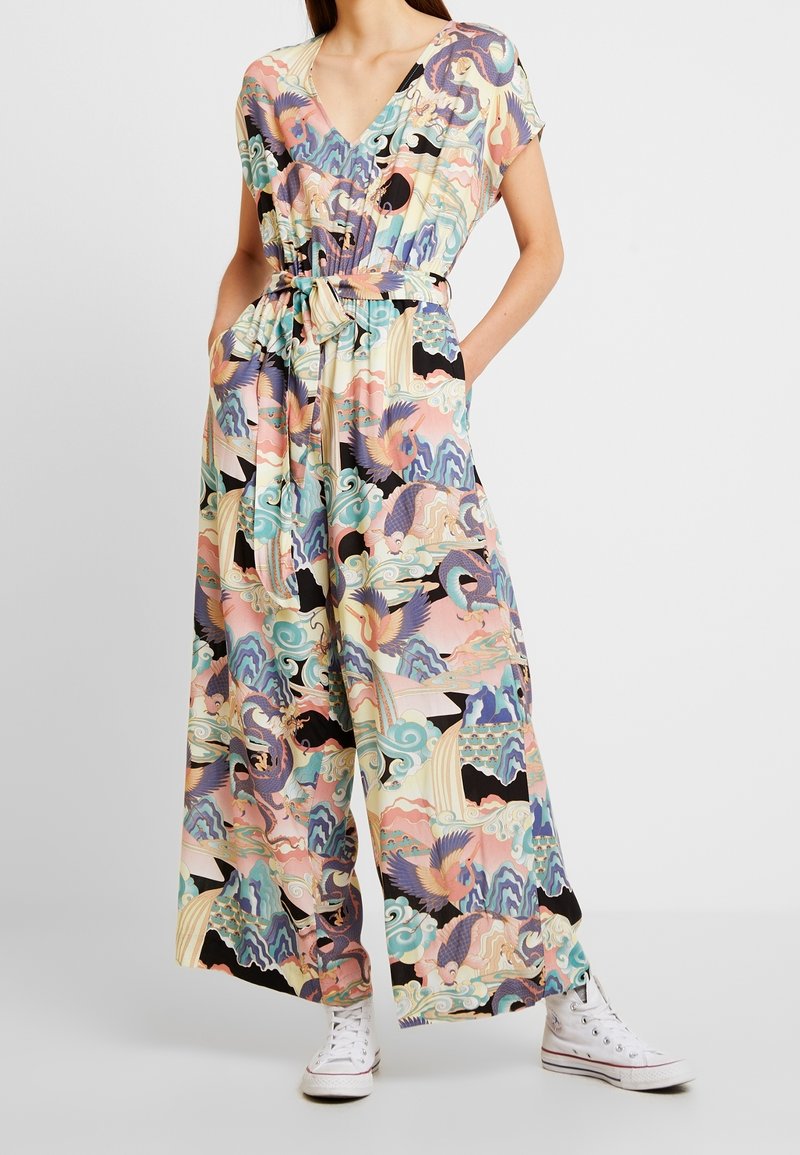 Woman wearing a colorful wide-leg jumpsuit with belt, featuring abstract bird and wave patterns, paired with white high-top sneakers.