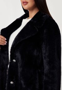 Cappotto in pelliccia sintetica nera con un ampio colletto e bottoni in tonalità argento, indossato sopra un maglione in cotone bianco, che mette in mostra una texture soffice e un design elegante.