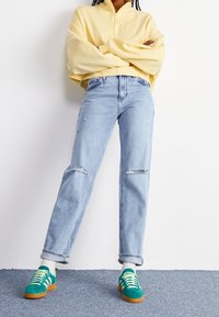 Keltainen vetoketjullinen collegepaita leveillä hihoilla; vaaleansiniset kulutetut farkut; vihreät ja oranssit nauhalliset tennarit valkoisilla sukilla.