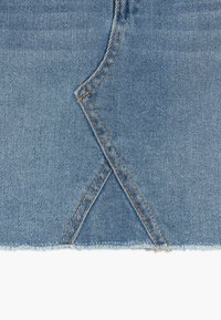 Detailní záběr modré džínoviny s diagonálním švem a oranžovým prošíváním, s raw, roztrženým dolním okrajem.