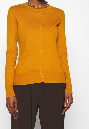 Personne portant un cardigan boutonné jaune moutarde et un pantalon marron foncé, accessoirisé avec une chaîne épaisse, sur fond blanc.