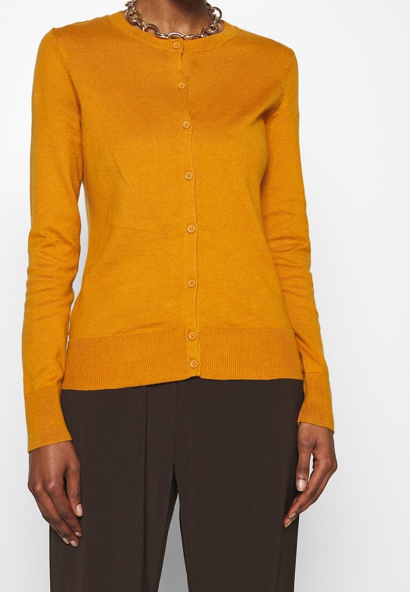 Personne portant un cardigan boutonné jaune moutarde et un pantalon marron foncé, accessoirisé avec une chaîne épaisse, sur fond blanc.
