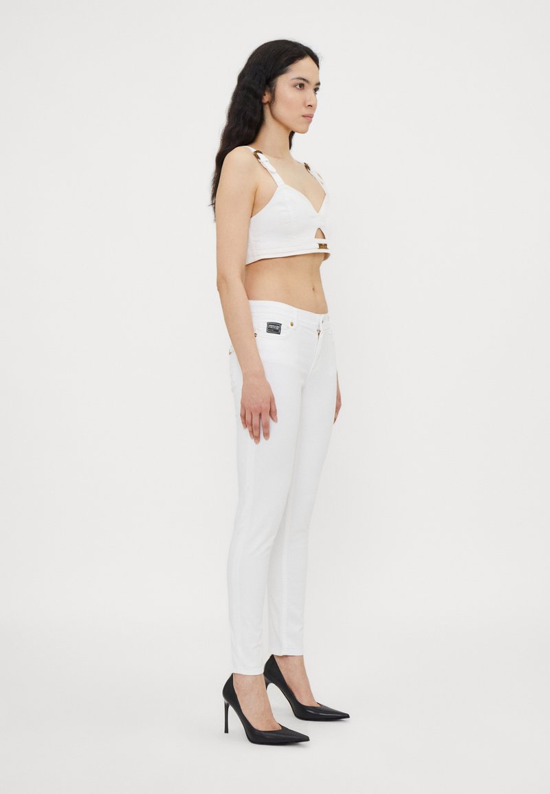 Witte cropped top met uitsnijding en gouden accenten, gecombineerd met aansluitende witte broek. Model draagt zwarte puntige hakken. Glad textuur door het hele outfit.