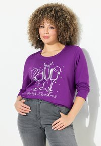 Maglietta lunga viola con grafica di bicchiere festivo e renna; presenta "Buon Natale" in bianco, abbinata a jeans grigi.