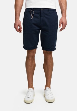 Mężczyzna ubrany w granatowe, podwinięte szorty ze splecionym sznurkiem, białe trampki i białą koszulkę, stojący na tle jednolitego tła.