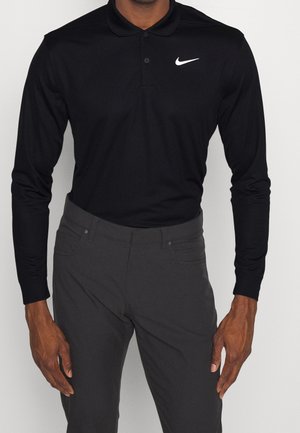 Mann trägt eng anliegendes schwarzes Nike-Poloshirt mit langen Ärmeln und dunkelgraue Hose, steht mit locker an den Seiten hängenden Händen.