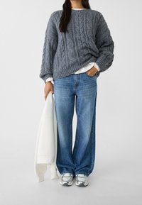 Grå kabelstickad tröja, lös passform, lager på lager över en vit skjorta. Blå vida jeans och metalliska sneakers fulländar outfiten.